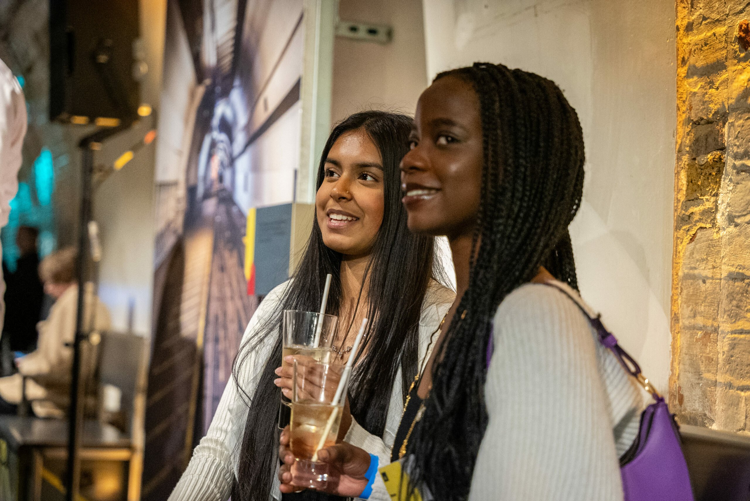 Two young women smiling and holding drinks at an indoor social event. One has long, straight dark hair and wears a white top, while the other has long braided hair, wears a light-colored sweater, and carries a purple handbag. They appear to be engaged in conversation, standing against a textured wall with warm lighting. A blurred background shows other attendees and a large printed image of a tunnel.