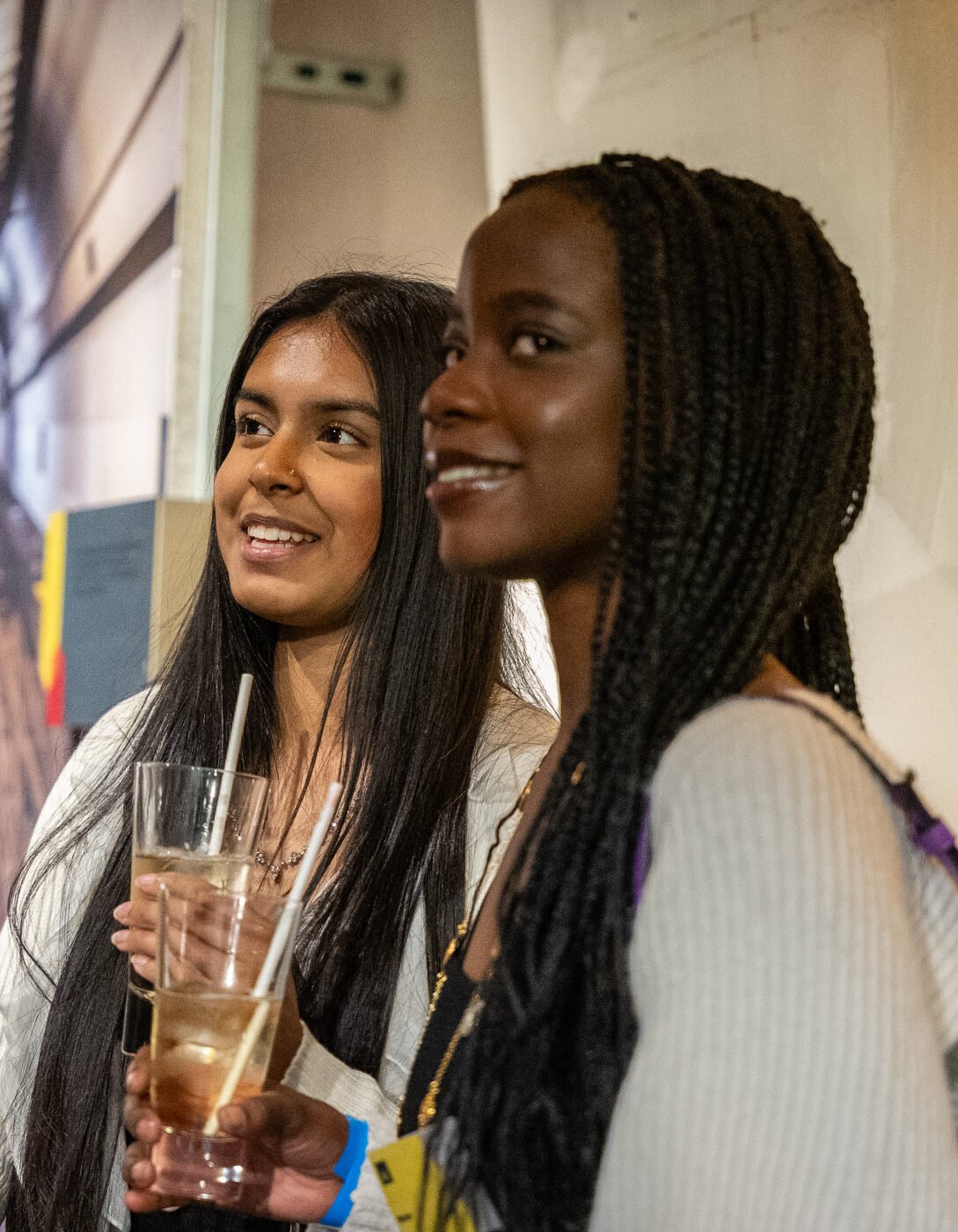 Two young women smiling and holding drinks at an indoor social event. One has long, straight dark hair and wears a white top, while the other has long braided hair, wears a light-colored sweater, and carries a purple handbag. They appear to be engaged in conversation, standing against a textured wall with warm lighting. A blurred background shows other attendees and a large printed image of a tunnel.