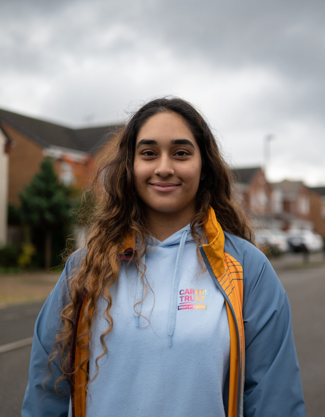 A young woman with long, wavy brown hair stands on a residential street, smiling gently at the camera. She wears a light blue hoodie with a 'Carers Trust' logo and a blue and yellow jacket. The background features suburban houses, parked cars, and an overcast sky.