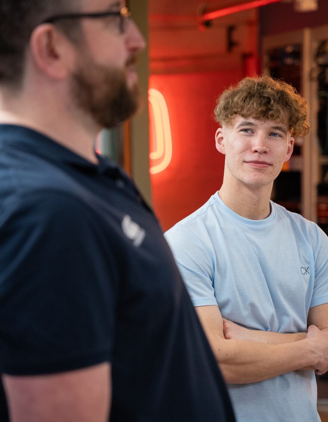 A young man with curly blond hair, wearing a light blue t-shirt, stands with his arms crossed in a modern workspace, looking attentively at an older man in the foreground who is wearing a navy polo shirt. The background features shelves with equipment, warm lighting, and a neon sign.