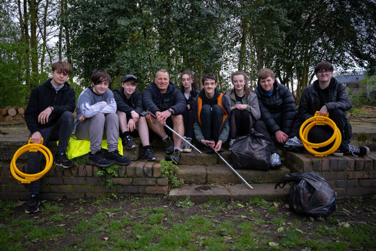 A group of young people sit together on outdoor stone steps in a wooded area, holding litter‑picking tools and bags filled with collected rubbish. Two individuals at each end hold yellow plastic hoops used for the activity. The group appears to be taking a break during an outdoor clean‑up session, with trees and greenery in the background.