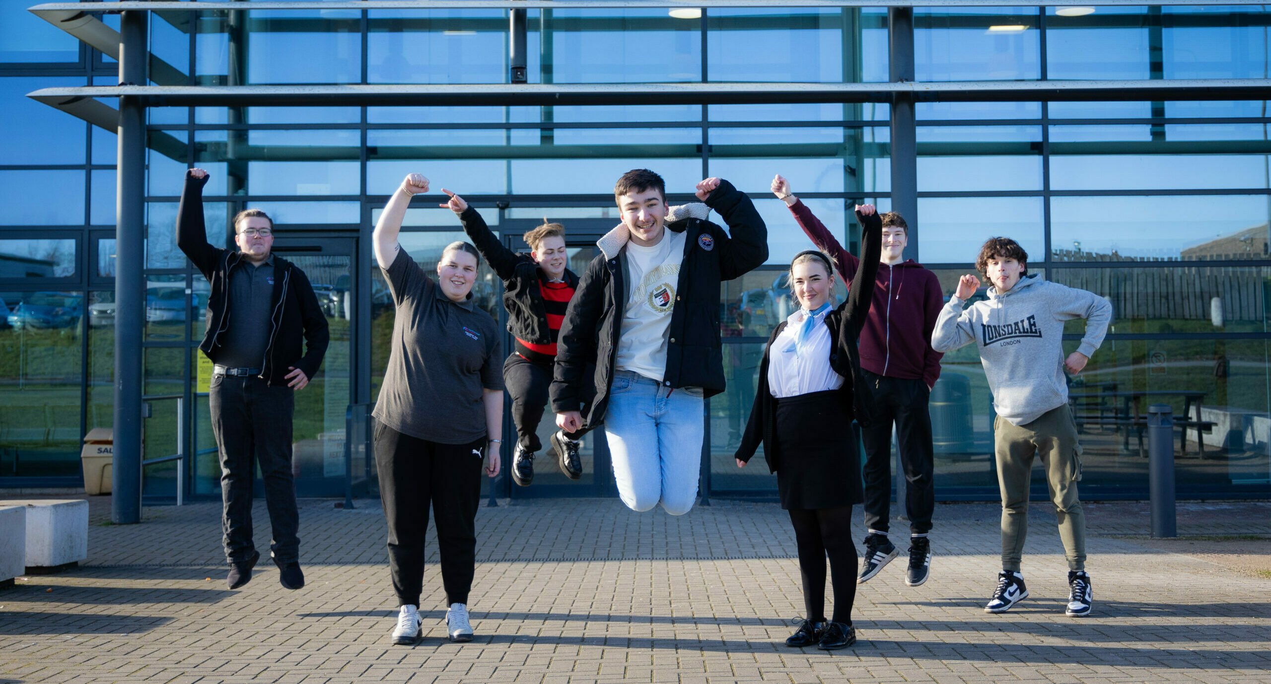 A group of eight young people is jumping in front of a modern glass building. They are all smiling and appear to be celebrating. The individuals are dressed in casual clothing, with some wearing jackets and others in hoodies. The background features a clear blue sky and the building's reflective glass facade.