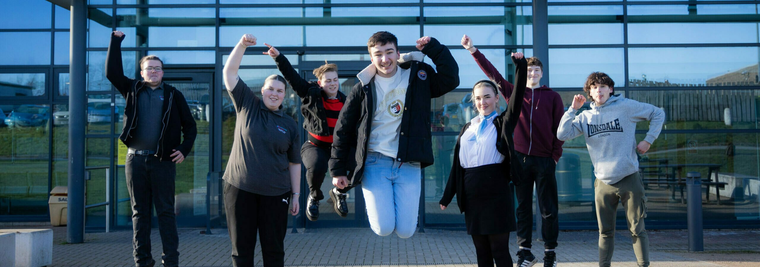 A group of eight young people is jumping in front of a modern glass building. They are all smiling and appear to be celebrating. The individuals are dressed in casual clothing, with some wearing jackets and others in hoodies. The background features a clear blue sky and the building's reflective glass facade.