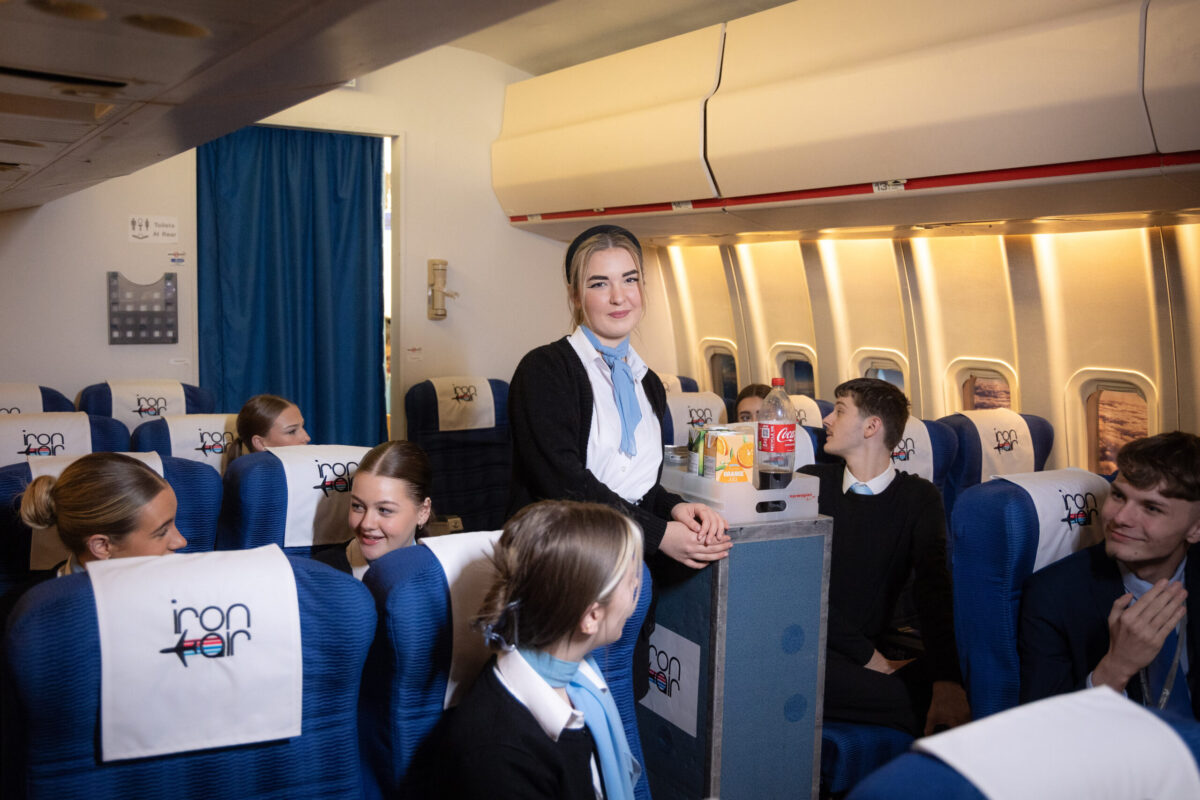 A student cabin crew member stands with a trolley in the aisle, there are other students sitting in the seats.