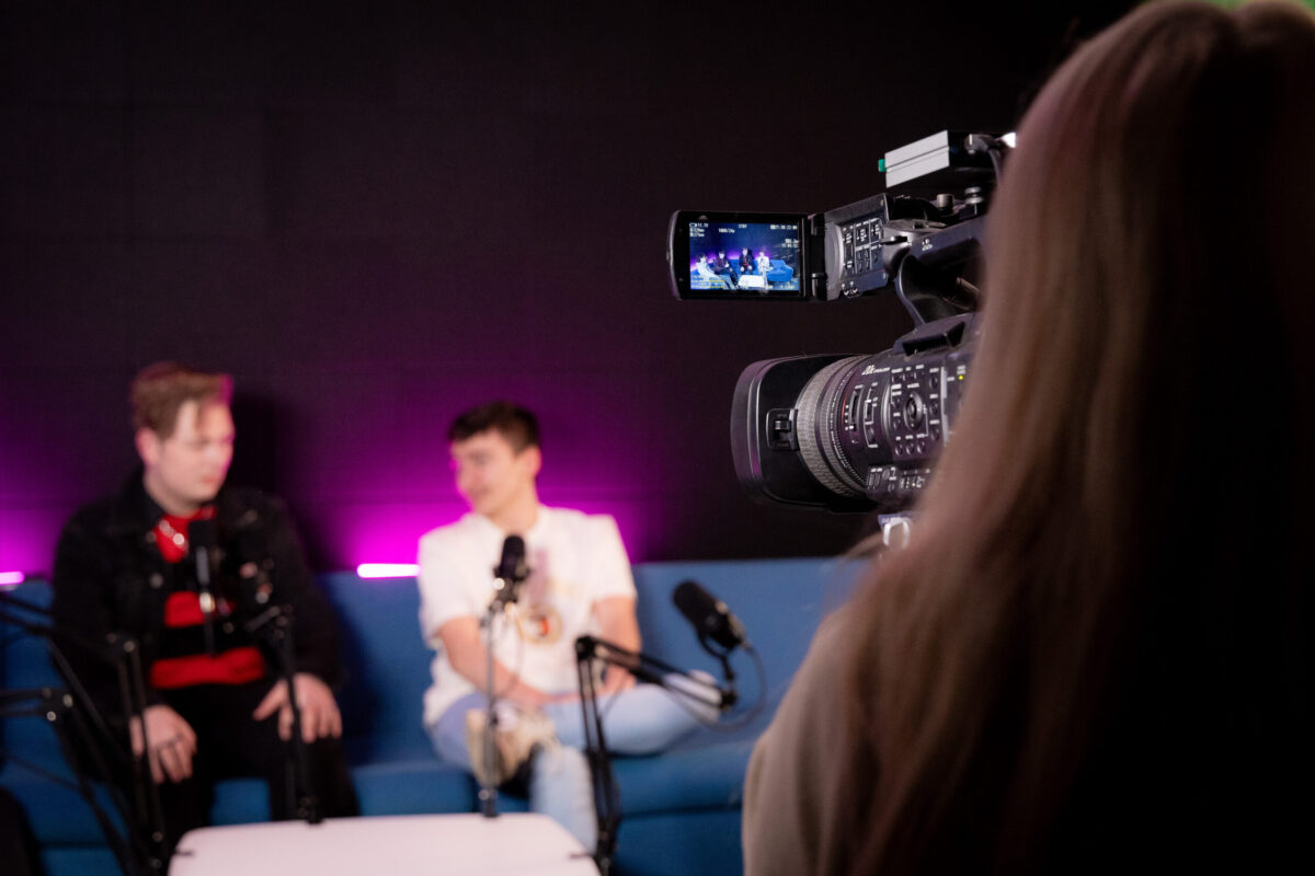 A person with a camera filming two people sitting on a sofa talking.