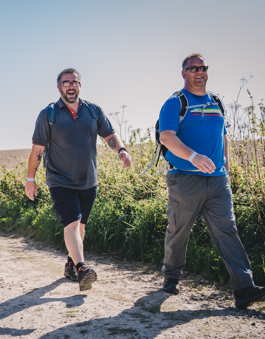Two men with backpacks are walking along a dirt path surrounded by greenery on a sunny day. The man on the left is wearing a dark polo shirt with orange accents, black shorts, hiking shoes, and glasses, and has a tattoo on his right arm. He is smiling broadly with his mouth open. The man on the right is wearing a bright blue t-shirt with a colorful stripe design, gray cargo pants, sunglasses, and a wristband. He is also smiling as they walk side by side. The background consists of tall grasses, wild plants, and a clear blue sky.