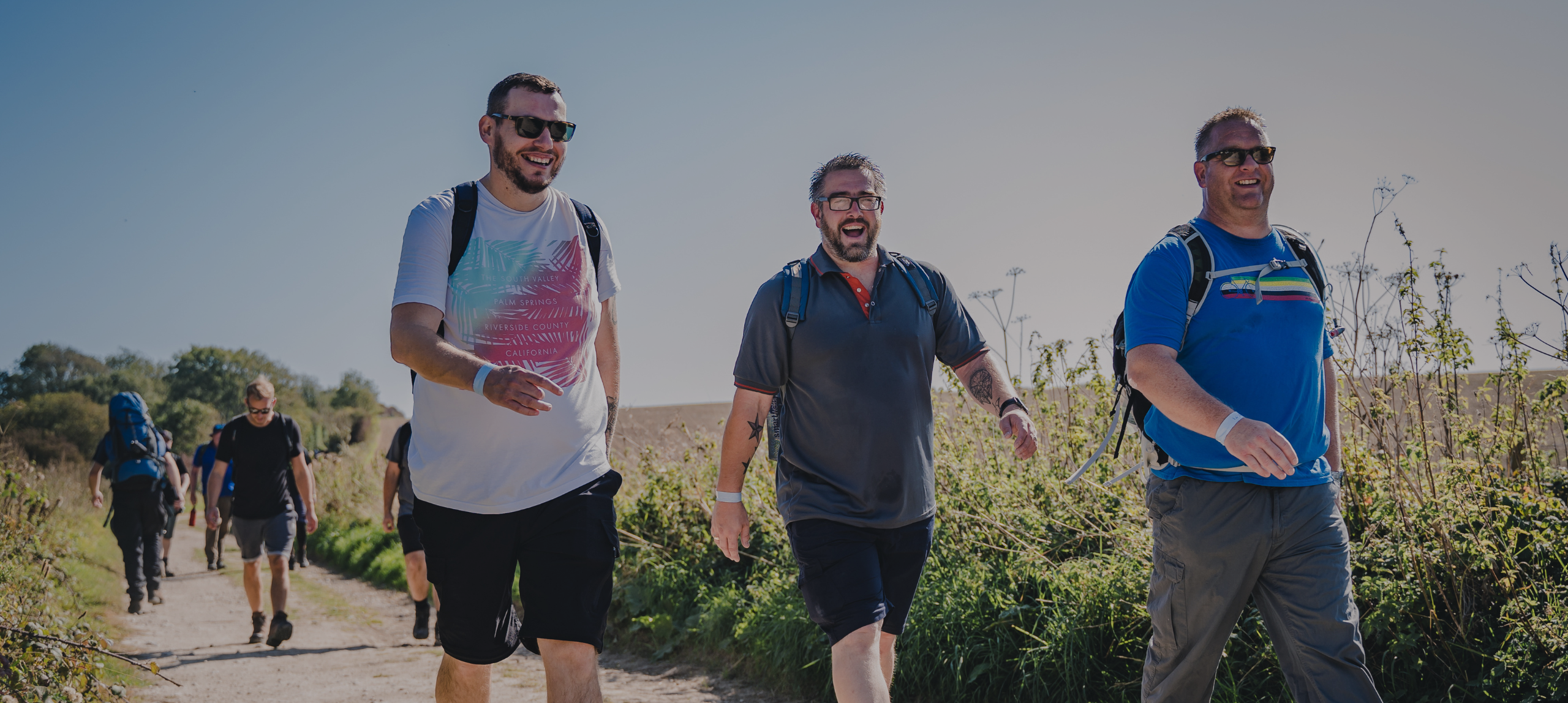 Three men with backpacks are walking along a dirt path surrounded by greenery on a sunny day. The man on the left is wearing a dark polo shirt with orange accents, black shorts, hiking shoes, and glasses, and has a tattoo on his right arm. He is smiling broadly with his mouth open. The man on the right is wearing a bright blue t-shirt with a colorful stripe design, gray cargo pants, sunglasses, and a wristband. He is also smiling as they walk side by side. The background consists of tall grasses, wild plants, and a clear blue sky.