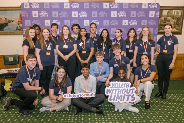 UK Youth Ambassadors wearing navy-blue polo shirts with the DofE logo and orange lanyards pose together in front of a purple 