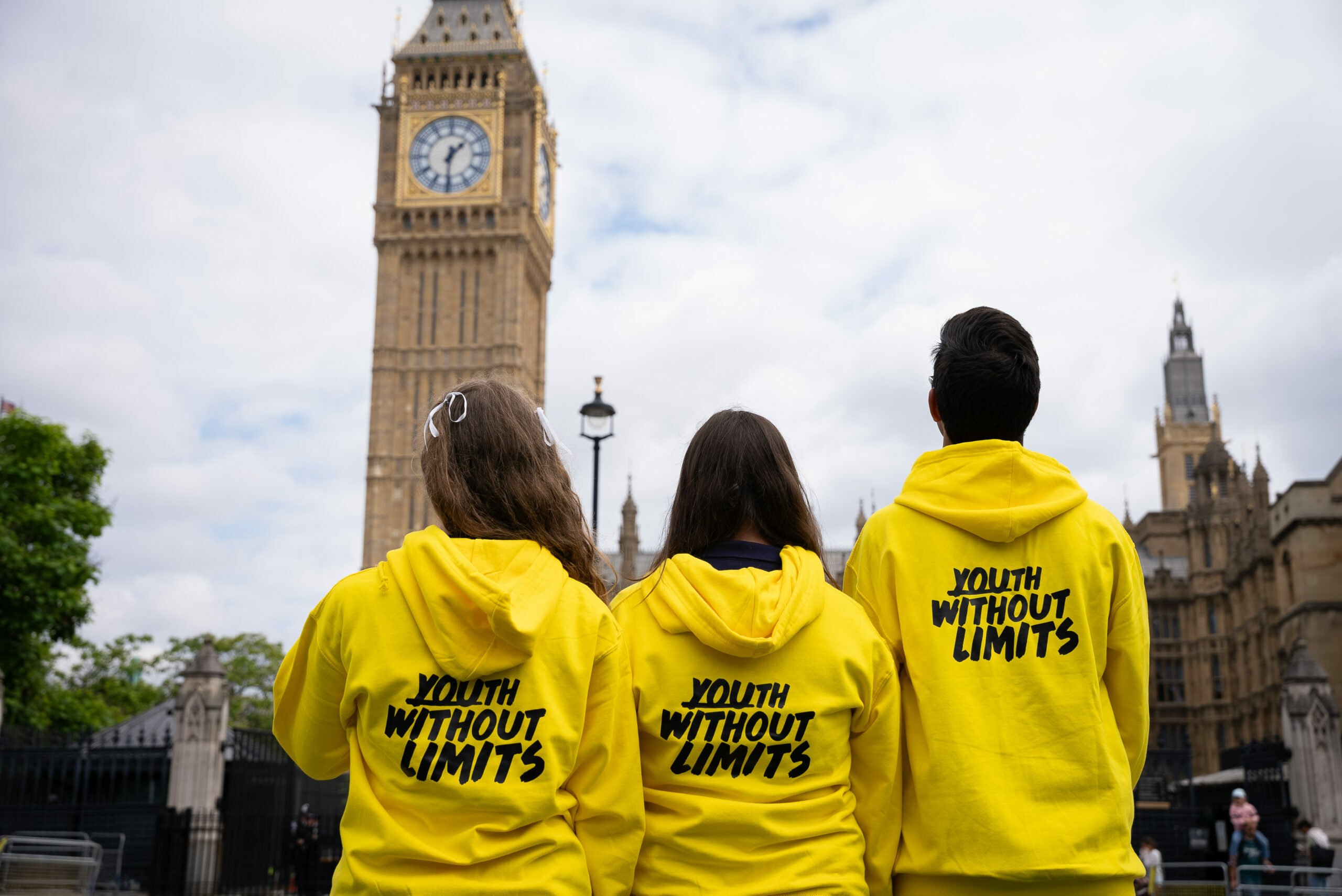 Three UK Youth Ambassadors stand with their backs to the camera, facing the Elizabeth Tower (Big Ben) in London. They are wearing matching bright yellow hoodies with the slogan 