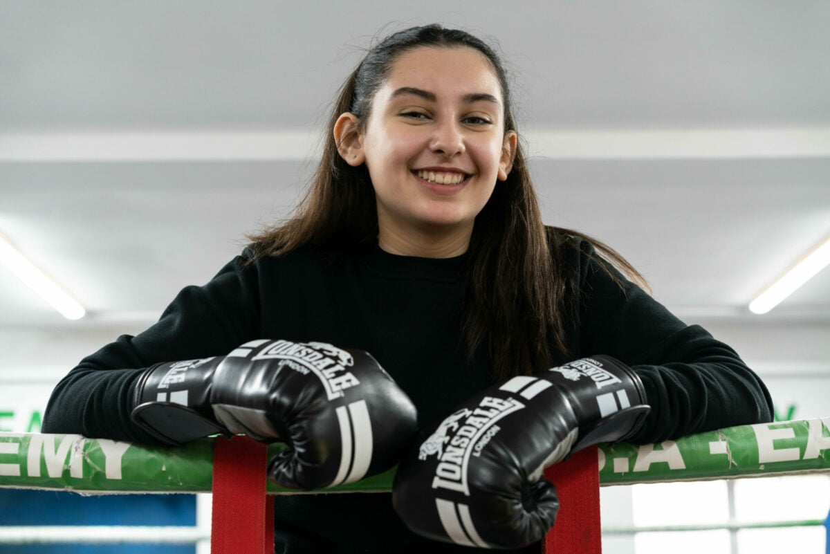 Summer is leaning on the ropes of a boxing ring, smiling confidently. She is wearing a black sweatshirt and black Lonsdale boxing gloves. The gym setting in the background is bright, with fluorescent lights overhead.