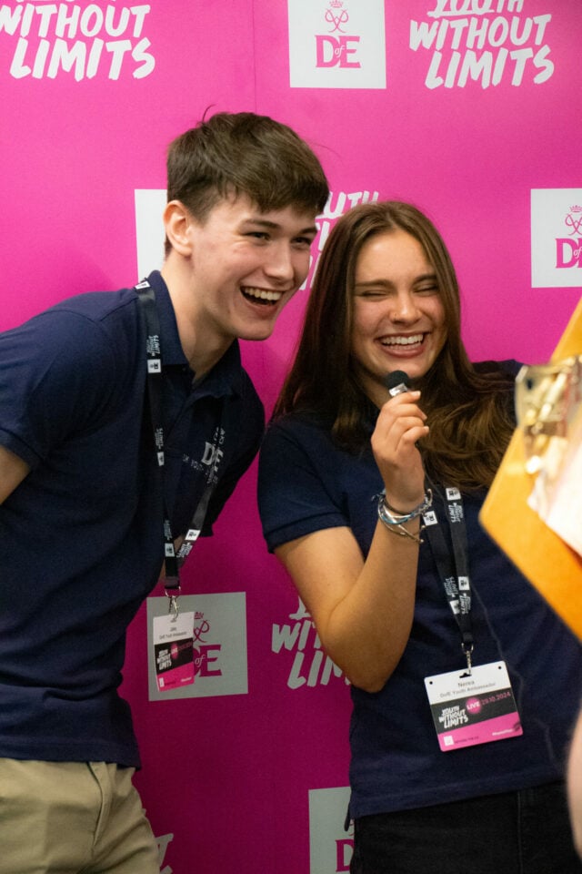 UK Youth Ambassadors wearing navy-blue DofE polo shirts and lanyards laugh together in front of a bright pink 