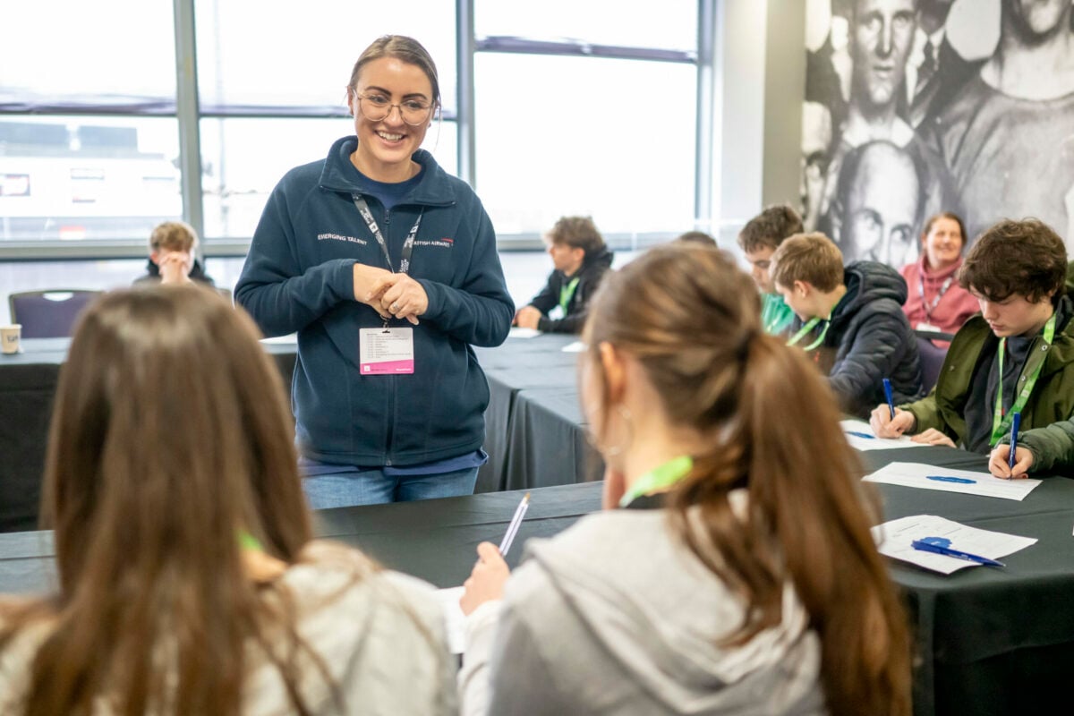 A smiling instructor stands in front of a group of students seated at tables in a classroom setting. The students, mostly teenagers, are engaged in writing on paper while listening attentively. The instructor is wearing a navy blue jacket and a name tag, and there are large windows in the background. The atmosphere appears collaborative and educational.