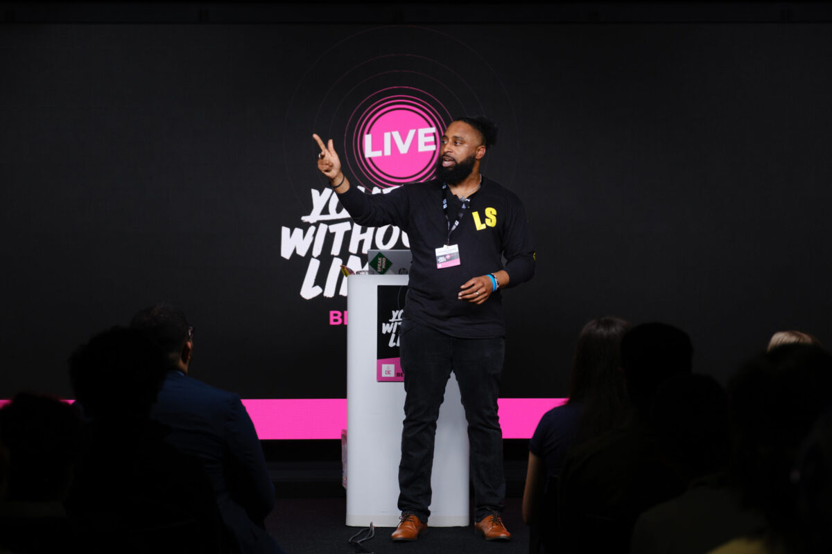 A speaker on stage at Youth Without Limits Live, his hand is in the air pointing upwards.