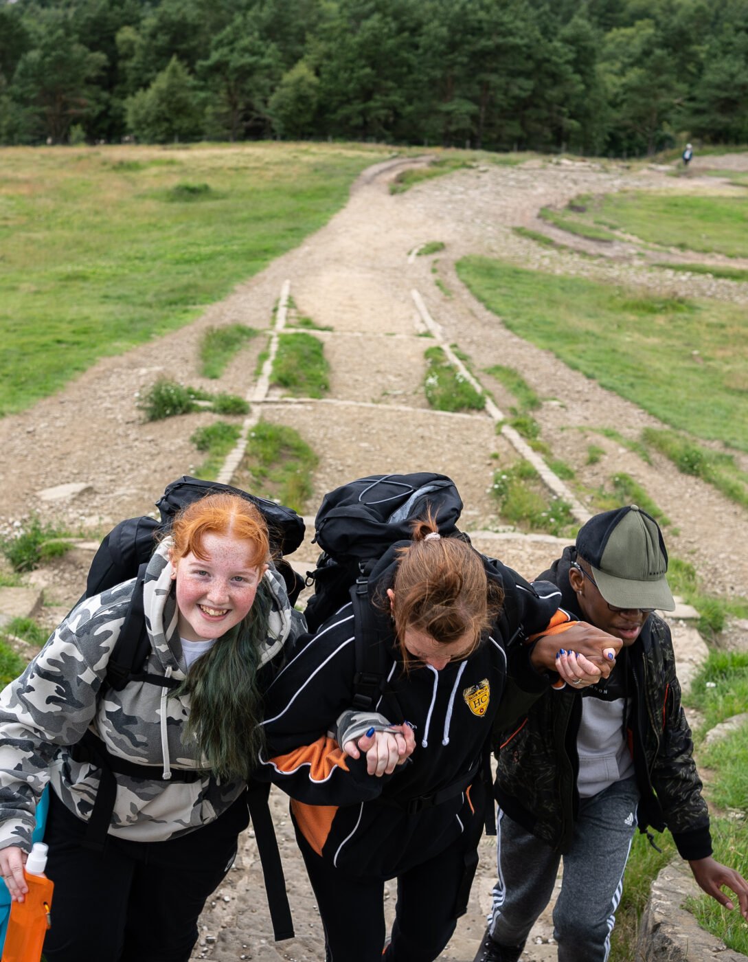 Three young people with backpacks are hiking up a rocky path in a green, open landscape. They are helping each other by holding hands for support, with one person in a camouflage hoodie smiling at the camera. The other two are focused on climbing, one wearing a black and orange jacket and the other in a cap and black jacket. The background features grassy fields, trees, and a wooden fence.