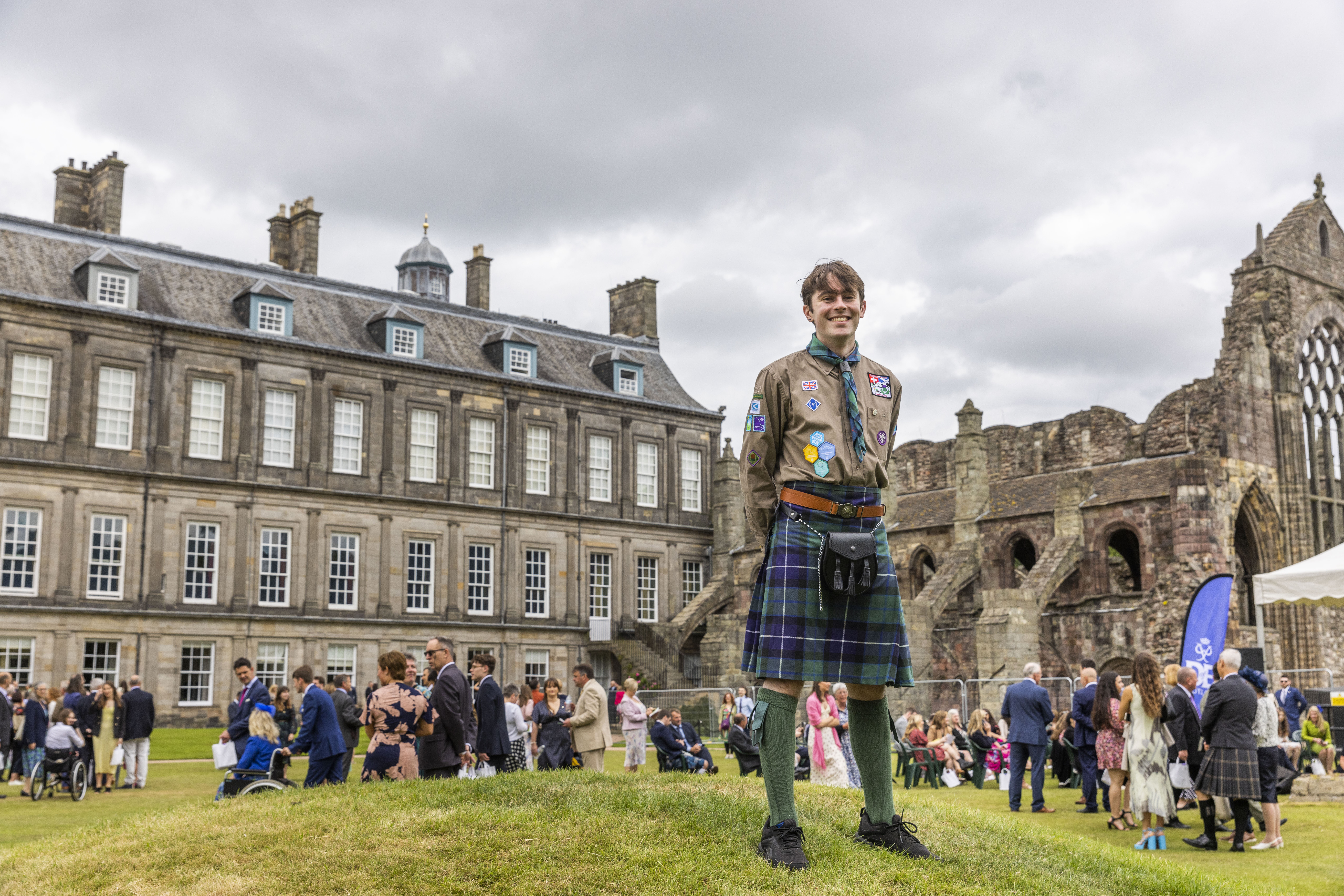 A young person wearing a kilt and Explorer Scout uniform standing in front of the Palace of Holyroodhouse.
