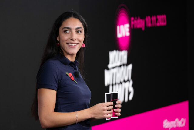 UK Youth Ambassador Lauren is standing on a stage, smiling as she looks to the side. She is wearing a navy-blue polo shirt with a red poppy pin and a headset microphone. She holds a small black notecard in her hands. Behind her, a large screen displays the words 