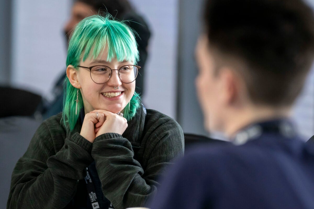 Youth Ambassador, Kea,engaging in conversation. They are wearing a dark sweater and have earrings. The background shows a blurred figure of another person, indicating a social setting.