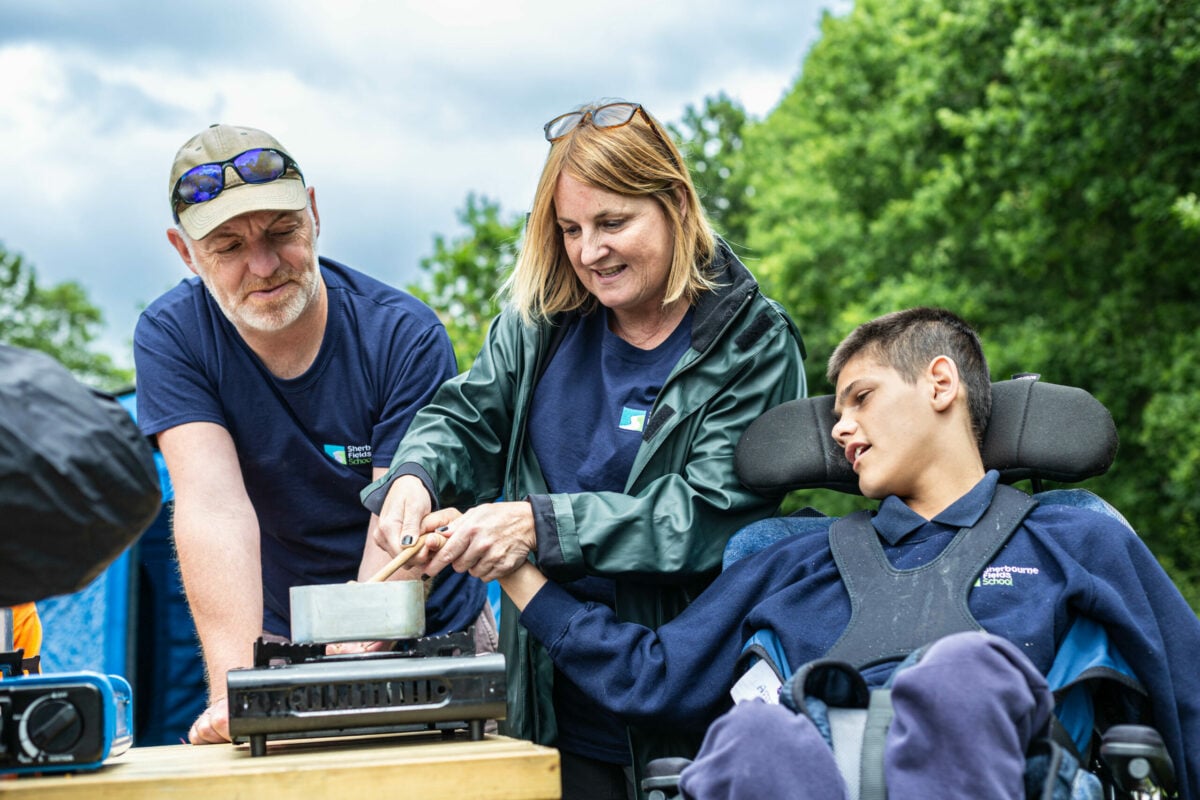 A man and a woman assist a young person in a wheelchair as they cook together outdoors. The woman is holding a pot over a portable stove, while the man supports the young person's arm. They are surrounded by greenery, and the sky is partly cloudy. All three are wearing casual clothing.