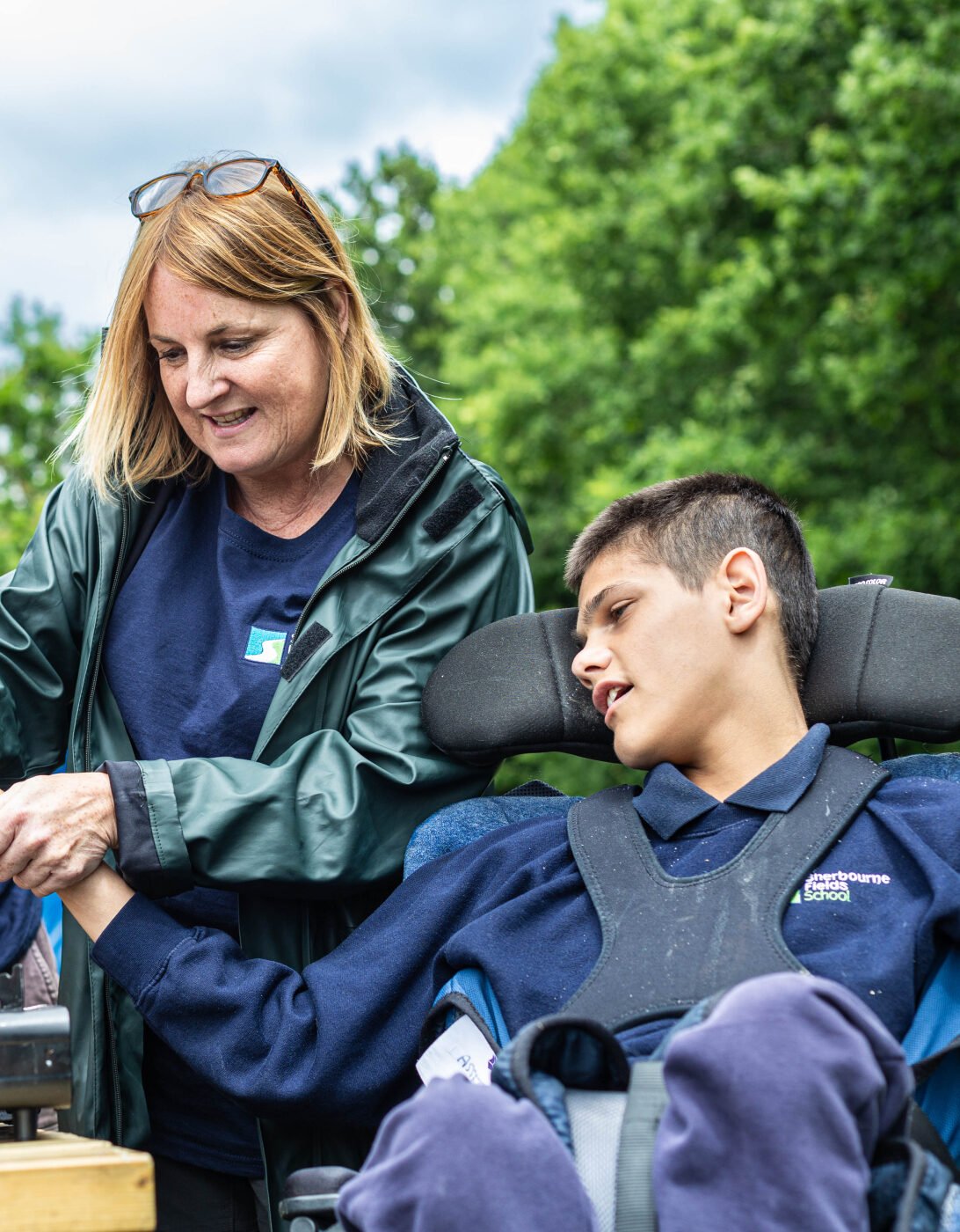 A man and a woman assist a young person in a wheelchair as they cook together outdoors. The woman is holding a pot over a portable stove, while the man supports the young person's arm. They are surrounded by greenery, and the sky is partly cloudy. All three are wearing casual clothing.