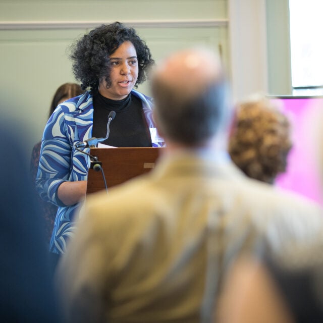 Lira stands at a podium, speaking into a microphone while holding a piece of paper. She is wearing a black top and a blue patterned blazer. The audience, seen from behind, listens attentively. The setting appears to be an indoor conference or formal event, with a large screen visible in the background.