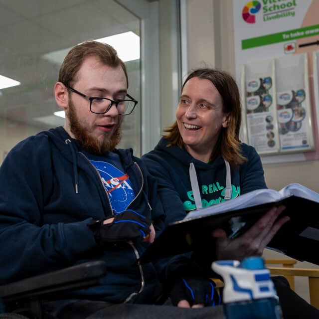 Brandon sits in a wheelchair, wearing a NASA t-shirt and a navy hoodie. He is holding a black folder and appears to be engaged in conversation. Beside him, DofE Leader, Ali is wearing a green hoodie, smiles warmly as she looks at him. They are sitting in what appears to be a school or learning center, with posters and brochures visible in the background.