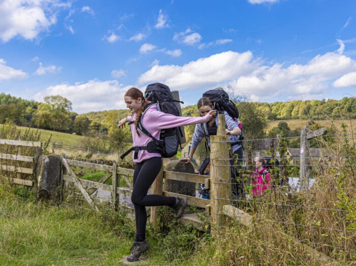 A young woman in a pink sweater and black leggings is climbing over a wooden fence while carrying a large backpack. In the background, two other individuals, also with backpacks, are navigating the fence. The scene is set in a lush, green landscape with trees and a blue sky dotted with clouds.