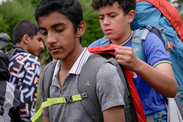 A group of young people wearing backpacks are outdoors, preparing for an expedition. The boy in the foreground, wearing a grey checkered shirt and a backpack with a chest strap, looks down with a focused expression. Behind him, another boy in a blue t-shirt is adjusting or securing his backpack. In the background, more participants with backpacks are visible, suggesting they are about to embark on a hike or outdoor adventure.