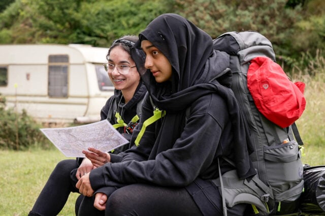 Two young women wearing black hoodies and backpacks are sitting outdoors in a grassy area. One of them, wearing a hijab, is attentively looking ahead, while the other, wearing glasses and a headband, is smiling and holding a map. A beige caravan is visible in the background, along with lush green trees and bushes.