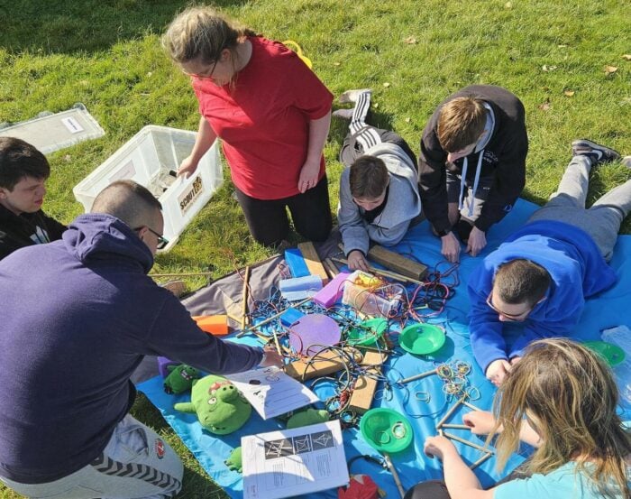 A group of six people is gathered on a blue tarp in a grassy outdoor setting. They are engaged in an activity involving various colorful materials, including wires, papers, and small green objects. Some individuals are sitting while others are kneeling, all focused on the task at hand. The scene is bright and sunny, with trees and a few people visible in the background.