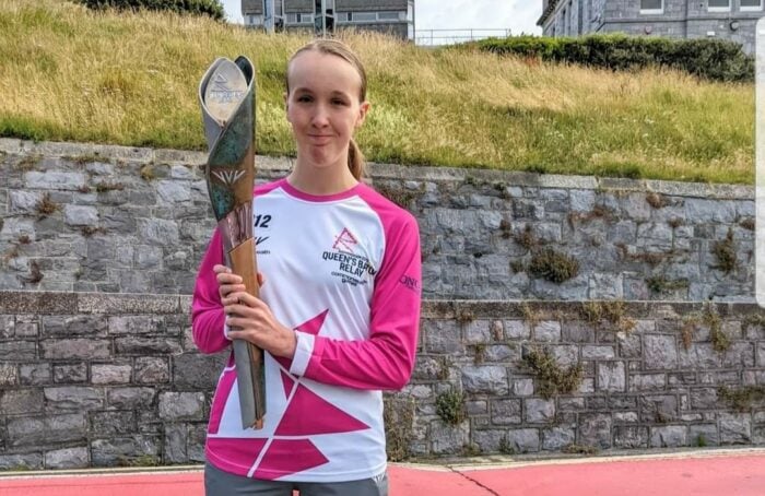 Abi stands on a running track, holding the Queen’s Baton from the Queen’s Baton Relay. She wears a pink and white relay shirt with the event logo and the number 12 printed on it. She has a proud and happy expression as she looks at the camera. Behind her is a stone wall with grass and buildings visible at the top.
