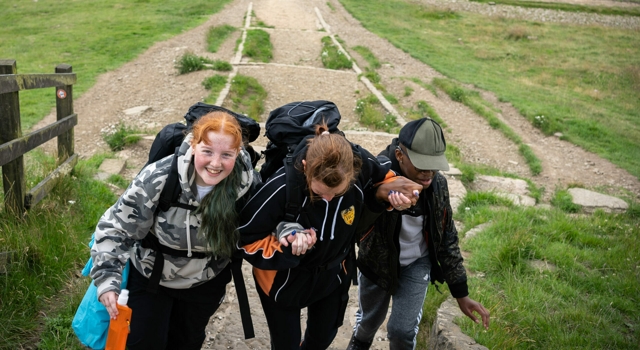 Three young people with backpacks are hiking up a rocky path in a green, open landscape. They are helping each other by holding hands for support, with one person in a camouflage hoodie smiling at the camera. The other two are focused on climbing, one wearing a black and orange jacket and the other in a cap and black jacket. The background features grassy fields, trees, and a wooden fence.