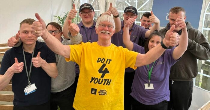 Eric in a bright yellow 'DO IT 4 YOUTH' T-shirt stands smiling at the front of a cheerful group, all giving thumbs up. The group, wearing matching purple uniforms and lanyards, pose enthusiastically inside a white marquee with draped fabric overhead and large windows in the background.