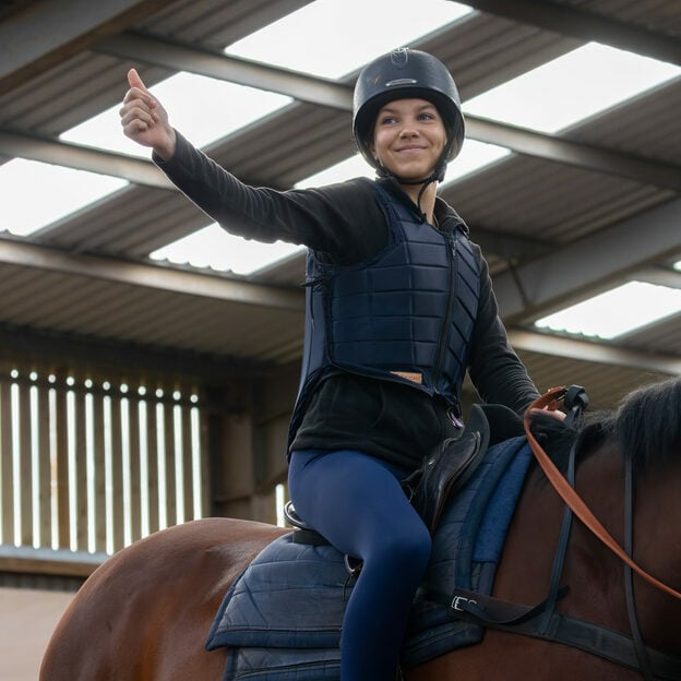 A young person riding a horse indoors giving a thumbs up.
