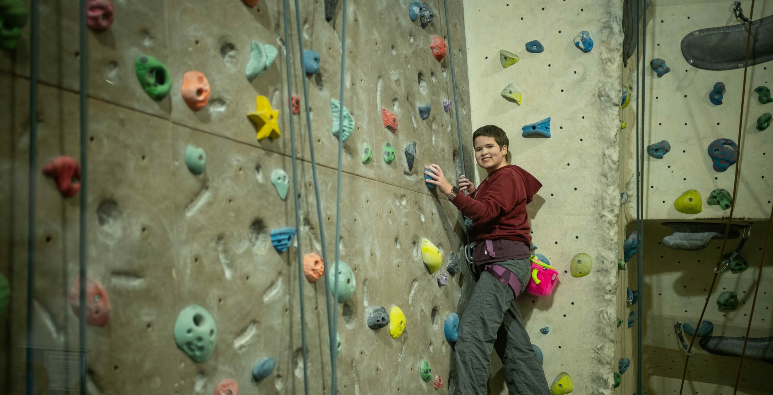 A person is climbing a colorful indoor rock climbing wall. They are wearing a maroon hoodie and gray climbing pants, with climbing shoes and a harness. The wall is adorned with various holds in different shapes and colors, and the climber is positioned mid-climb, smiling confidently.