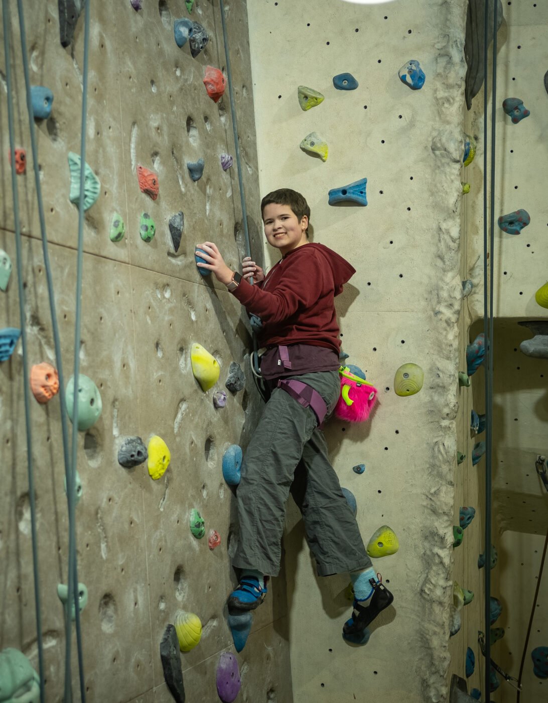 A person is climbing a colorful indoor rock climbing wall. They are wearing a maroon hoodie and gray climbing pants, with climbing shoes and a harness. The wall is adorned with various holds in different shapes and colors, and the climber is positioned mid-climb, smiling confidently.