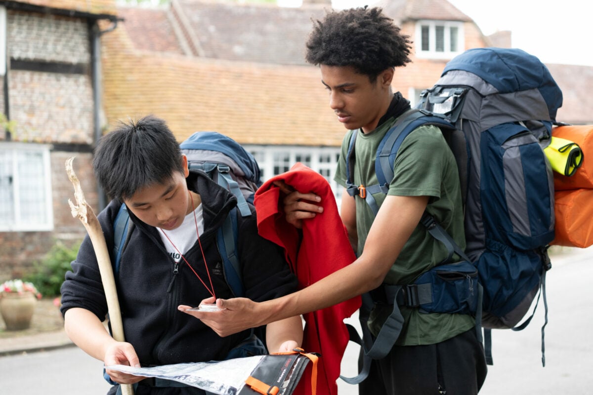 Two young men are outdoors, preparing for an adventure. One is wearing glasses and a white shirt, holding a map or a device, while the other, dressed in a black hoodie, is examining a walking stick. Both have backpacks on, and they appear focused on their task. In the background, there are white and brick buildings.