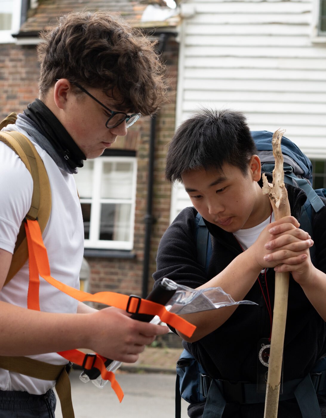 Two young men are outdoors, preparing for an adventure. One is wearing glasses and a white shirt, holding a map or a device, while the other, dressed in a black hoodie, is examining a walking stick. Both have backpacks on, and they appear focused on their task. In the background, there are white and brick buildings.
