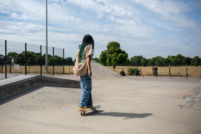 Amaris rides a skateboard at an outdoor skate park on a sunny day. They are wearing a beige plaid shirt, loose blue jeans, and orange sneakers. The skate park has ramps with graffiti, and a grassy field with trees is visible in the background beyond a metal fence.
