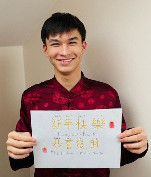 Evan is wearing a red, traditional Chinese shirt, holding a poster that says Happy Lunar New Year in Chinese.