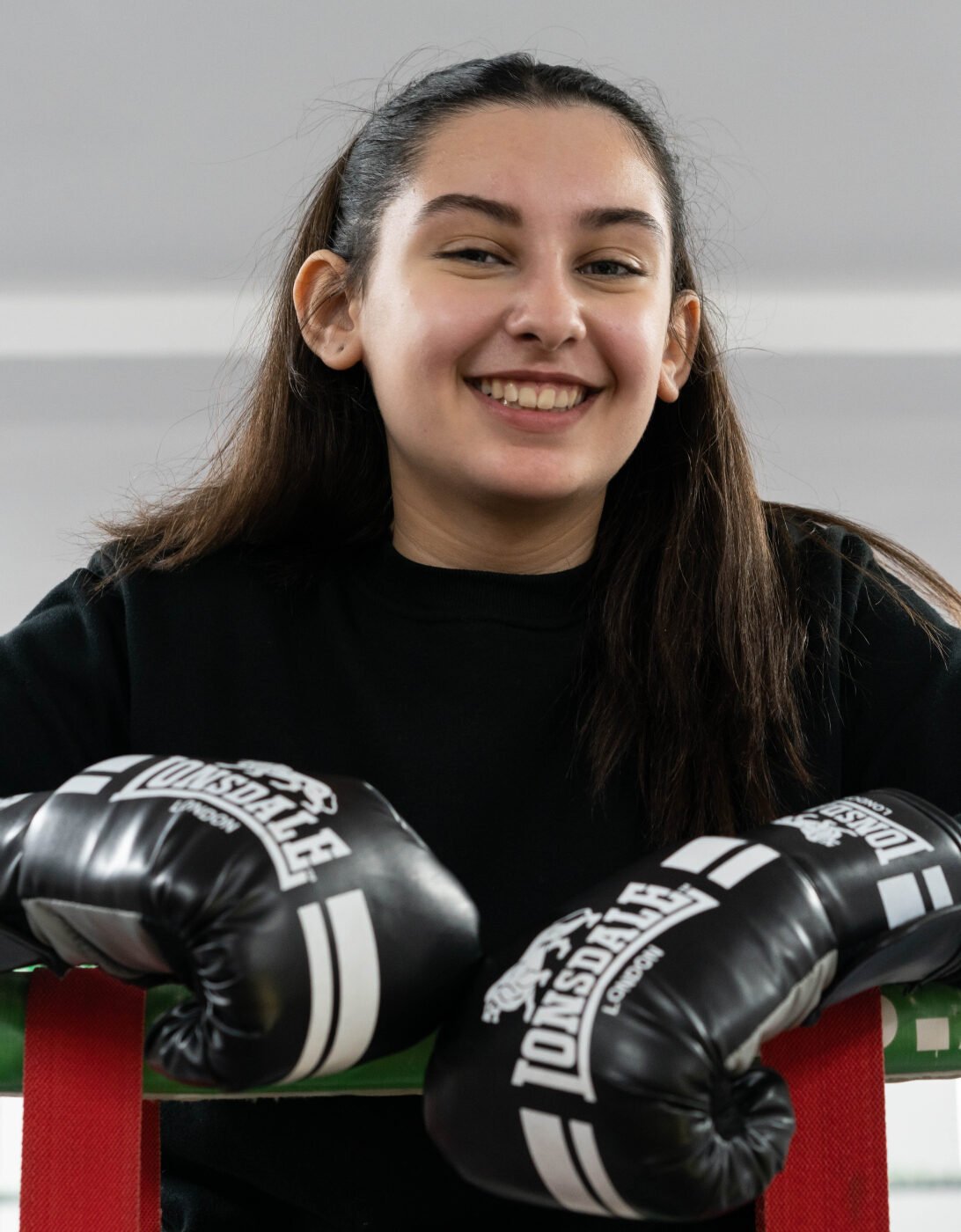 Summer is leaning on the ropes of a boxing ring, smiling confidently. She is wearing a black sweatshirt and black Lonsdale boxing gloves. The gym setting in the background is bright, with fluorescent lights overhead.