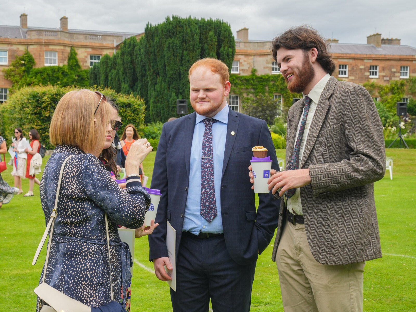 Three people in suits and a dress are chatting in Hillsborough Palace Gardens.