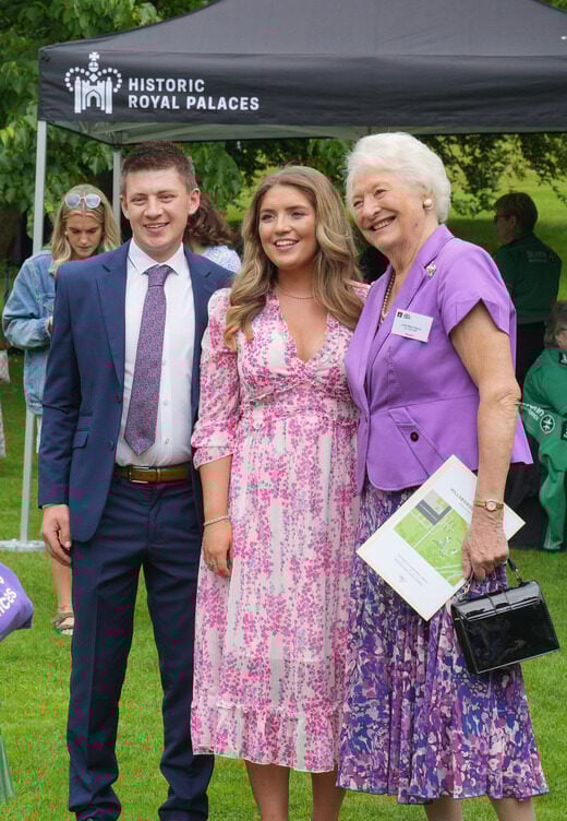 Three people dressed in a suit and nice dresses stand together and smile in Hillsborough Palace Gardens.