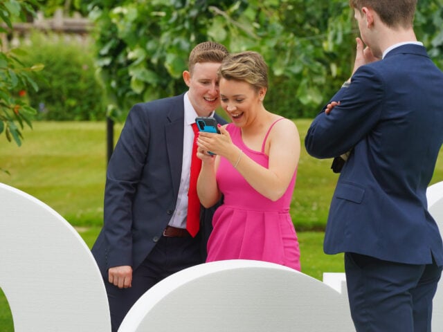 Two people in a suit and a pink dress smile and look at a phone, a person in a suit looks at them.