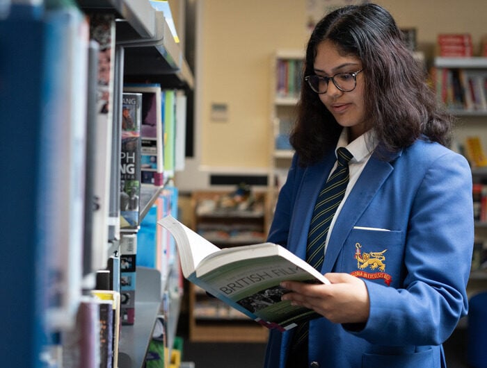 A young person in a blue school uniform reading a book standing next to a library shelf.