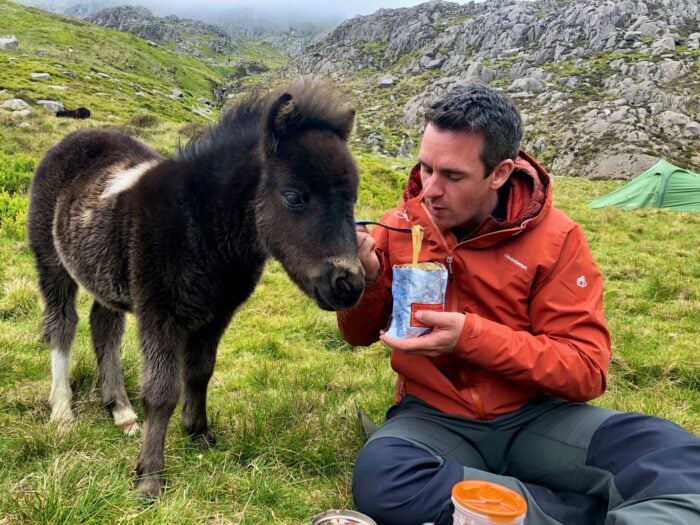 David Love, dressed in an orange outdoor jacket and sitting on grassy terrain in a mountainous area, shares his meal with a curious, fluffy black and white pony. A green tent is visible in the background among rocky hills and misty peaks, suggesting a camping trip in a remote, scenic location.