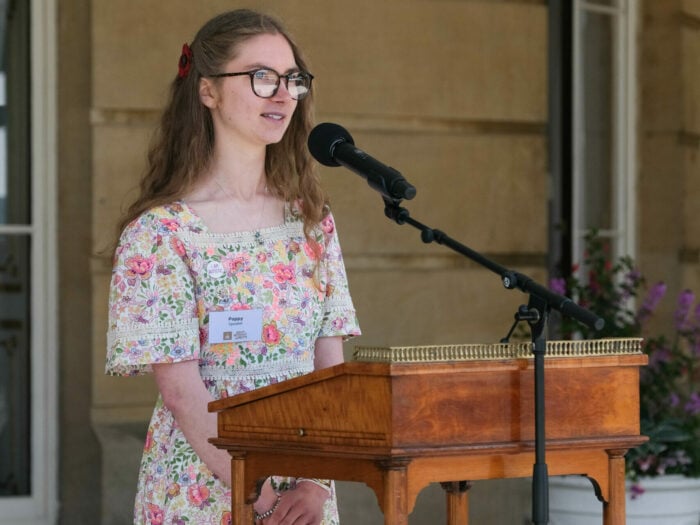Poppy stands at a wooden podium with a microphone, speaking confidently. She is wearing glasses and a light-colored dress with a floral pattern. The background shows part of a grand building with stone walls and potted flowers, suggesting a formal outdoor setting.
