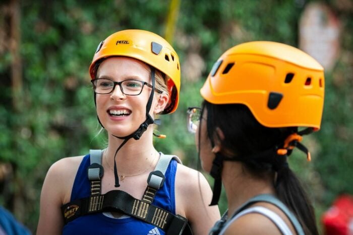 Two young women are wearing bright orange climbing helmets and harnesses, smiling and engaging in conversation. The background features greenery, suggesting an outdoor climbing or adventure setting. One woman has glasses and is wearing a blue tank top, while the other has long hair tied back.