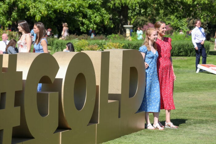 Two women pose beside a large gold hashtag sculpture reading '#GOLD' in a sunny garden setting, surrounded by other people enjoying the event. One wears a blue floral dress and the other a red floral dress.
