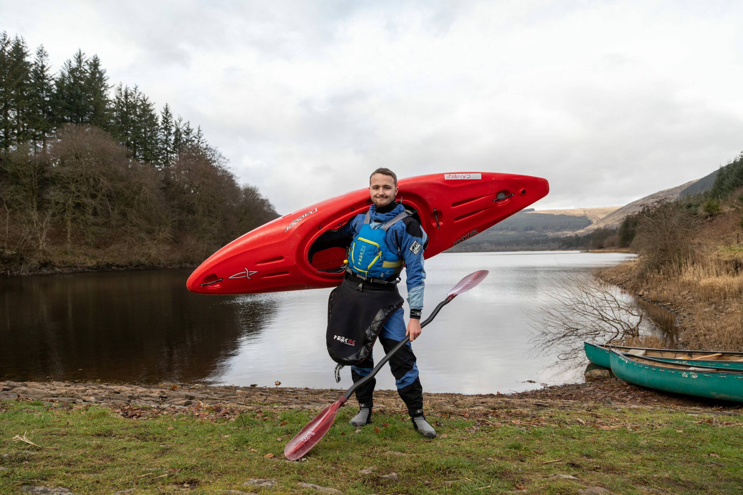 Elis-Llyr, wearing a blue kayaking drysuit and safety gear, stands on the shore of a lake carrying a red kayak on his shoulder and holding a paddle. The lake is surrounded by trees and hills under a cloudy sky, with a green canoe resting on the shore nearby.