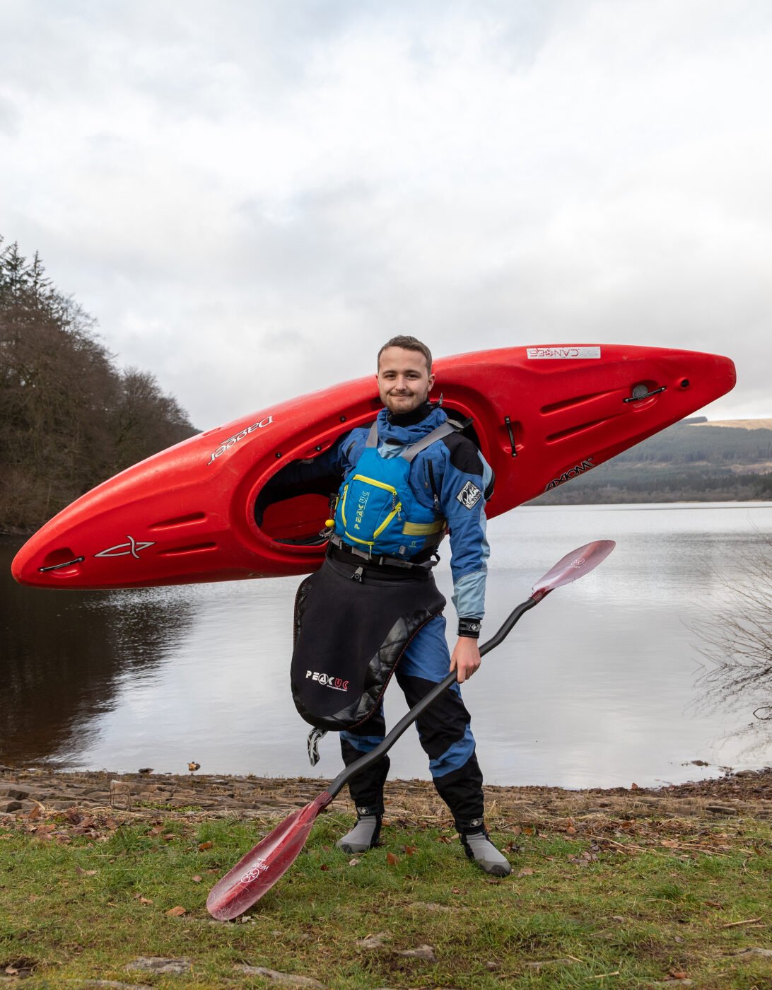 Elis-Llyr, wearing a blue kayaking drysuit and safety gear, stands on the shore of a lake carrying a red kayak on his shoulder and holding a paddle. The lake is surrounded by trees and hills under a cloudy sky, with a green canoe resting on the shore nearby.
